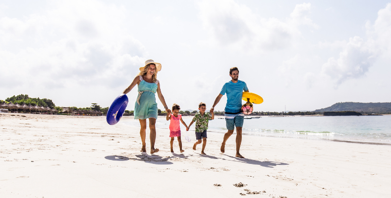 A couple enjoying travel during pregnancy and holding hands with their two children on the beach.