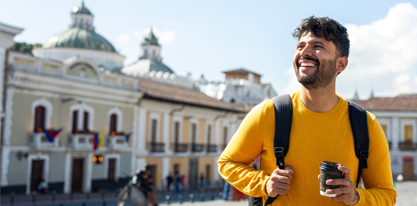 Young man smiling with a backpack and holding coffee in front of San Francisco church in Quito, Ecuador