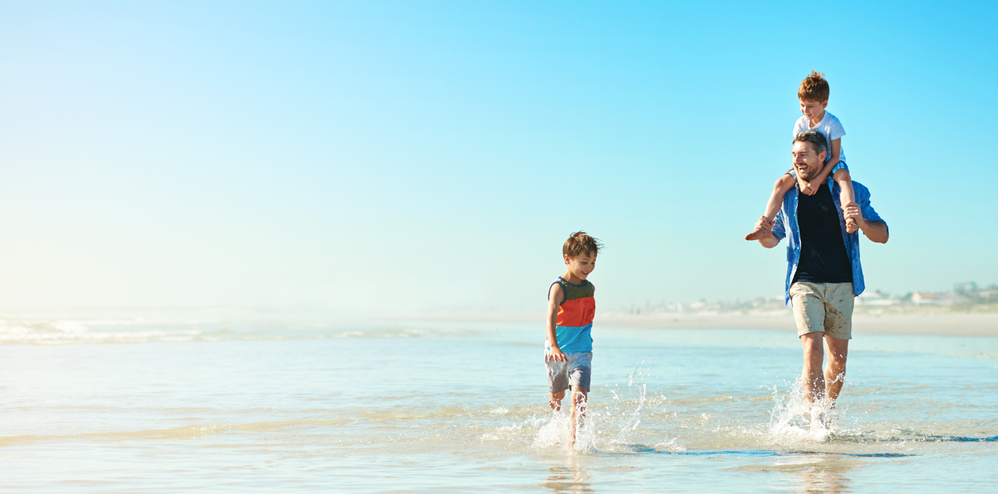 Father walking on the beach with his son on his shoulders and another alongside him with long term travel insurance