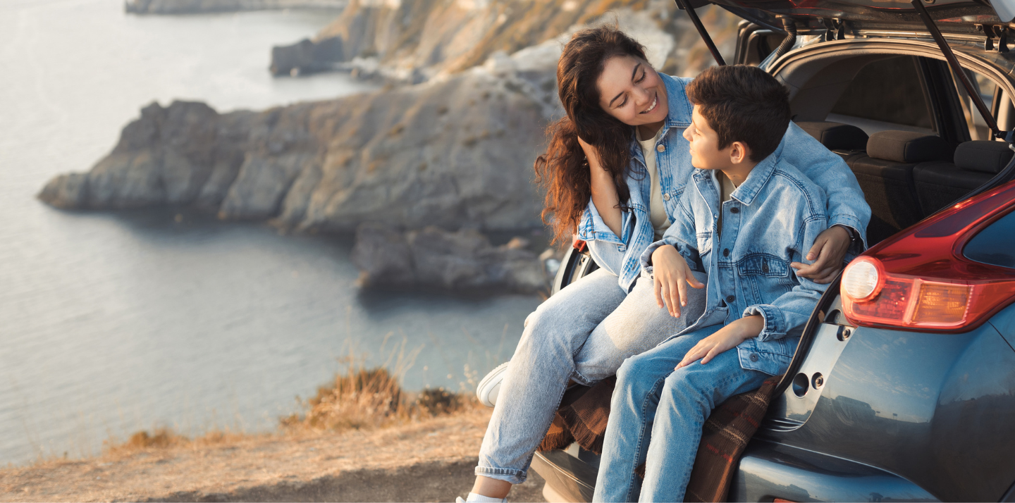 Mother with her arm around her son in the car trunk on a cliff overlooking the water with road trip travel insurance