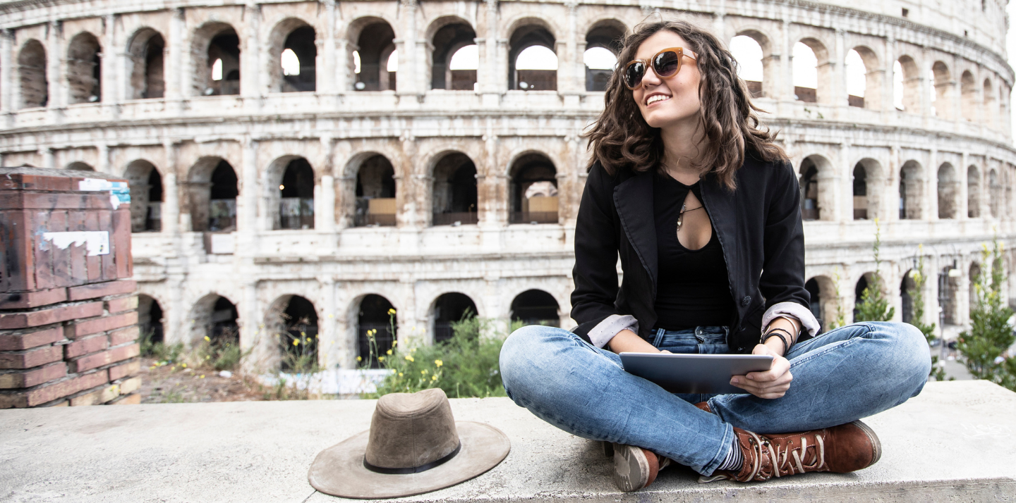 Young woman sitting with crossed legs and holding a tablet in front of the Colosseum