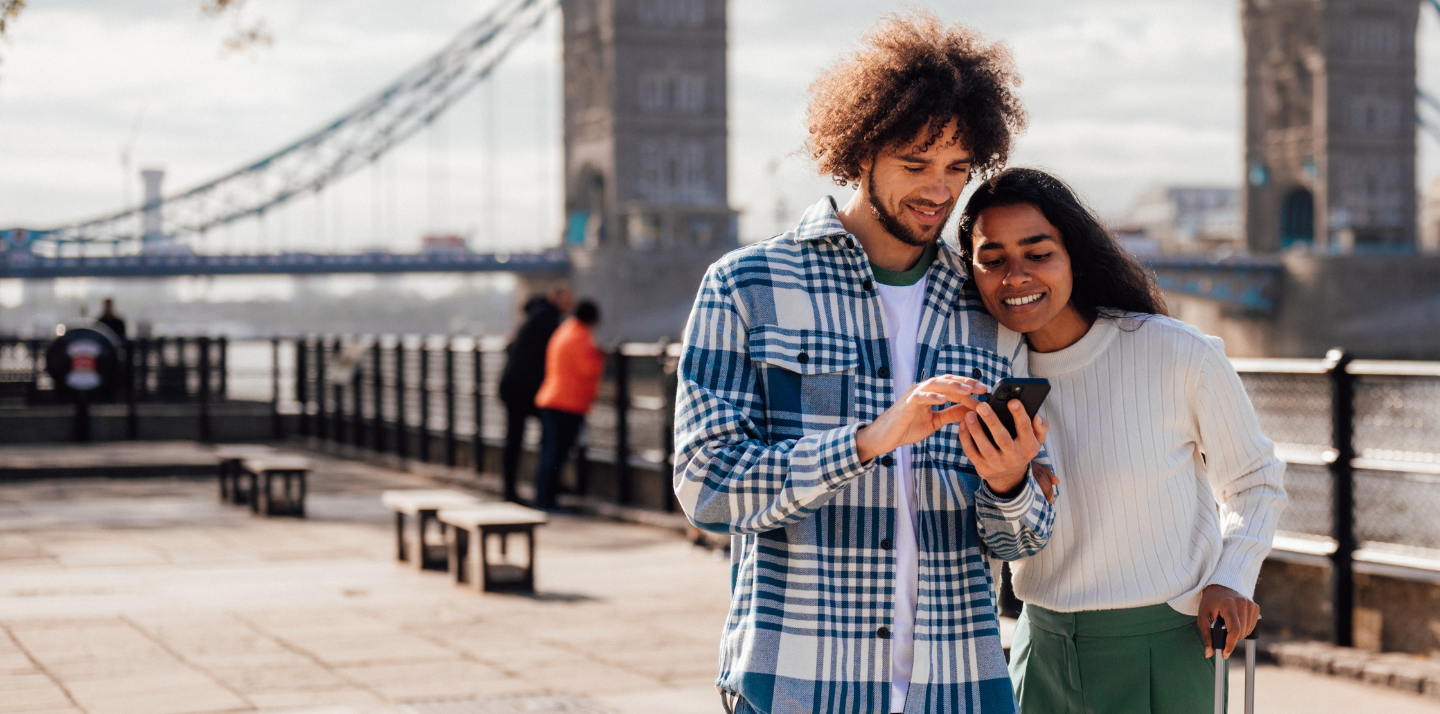 Couple in the U.K. in front of Tower Bridge smiling and looking at the Travelex Travel On app on their phone