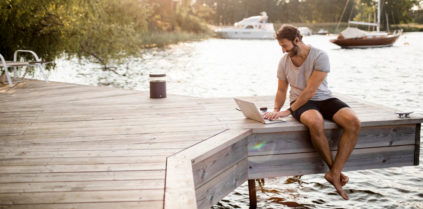 Young man sitting on a dock over water looking at his laptop and smiling