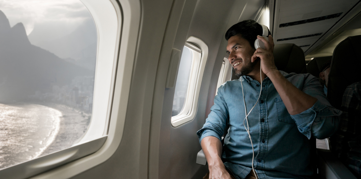 Young smiling man looking out the window of an airplane with headphones on and flight travel insurance