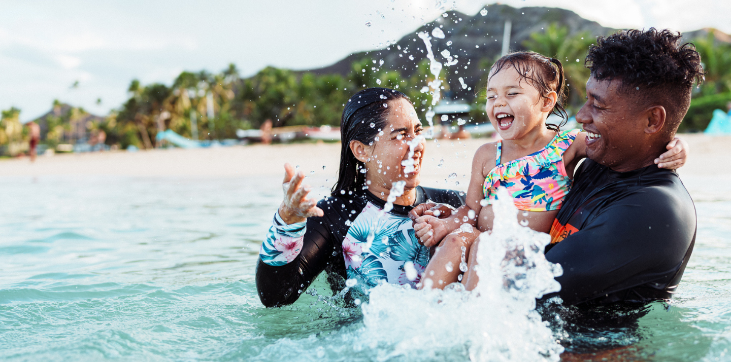 Multiracial couple with young daughter splashing in the ocean and smiling while traveling with travel insurance benefits