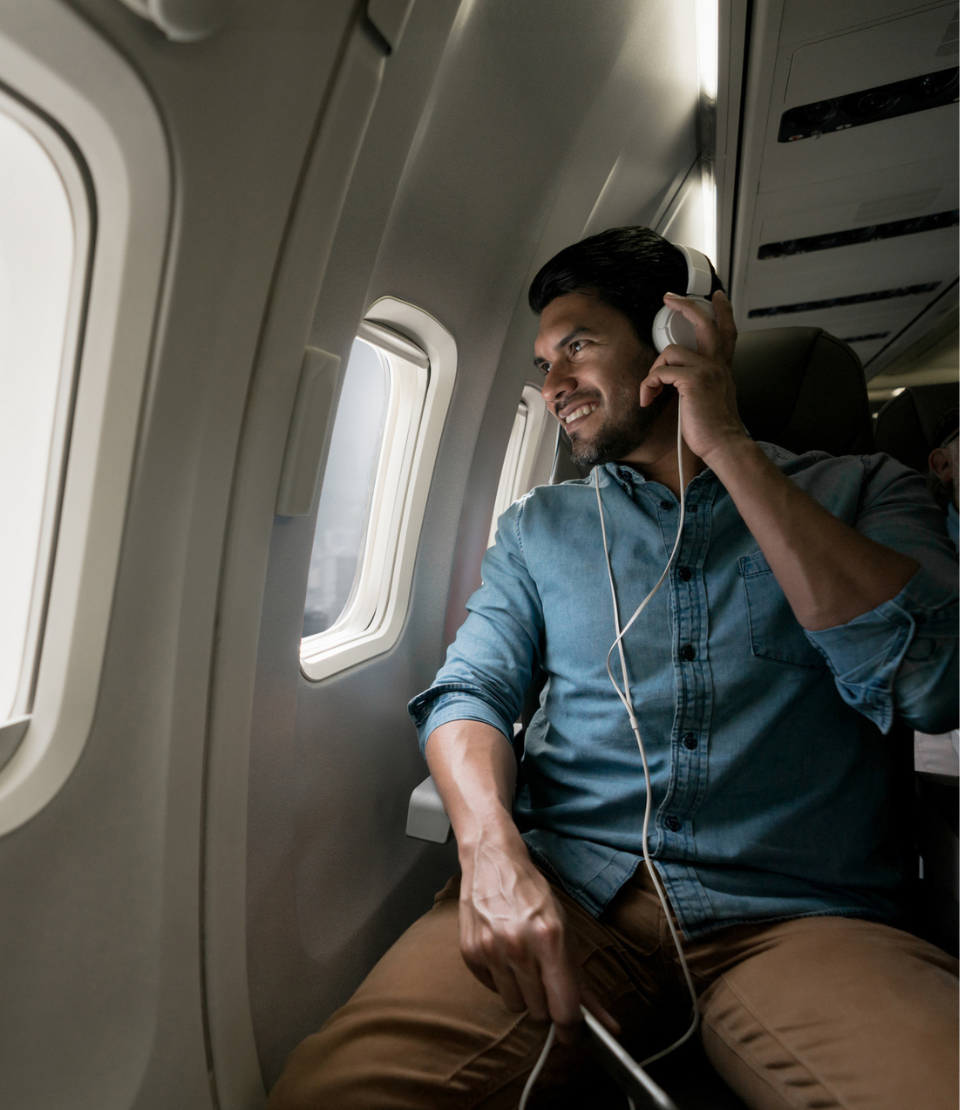 Young smiling man looking out the window of an airplane with headphones on and flight travel insurance