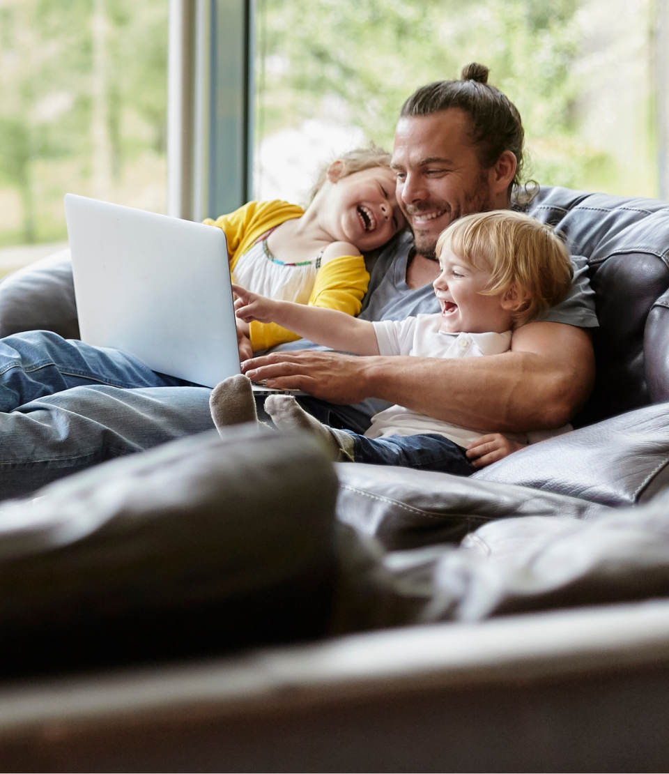 Father smiling with two young children on the couch in front of a laptop with travel insurance for trip cancellation