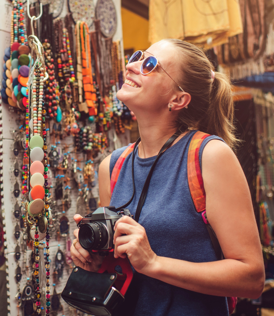 Young woman smiling looking at a store in a marketplace with her camera and solo travel insurance