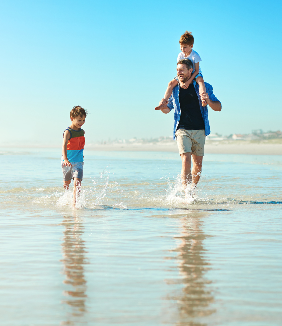 Father walking on the beach with his son on his shoulders and another alongside him with long term travel insurance