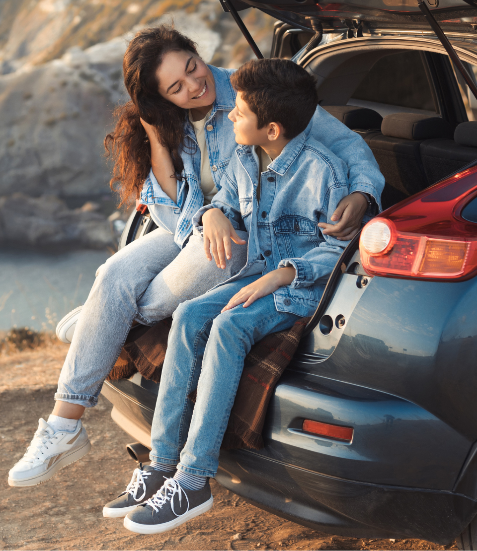 Mother with her arm around her son in the car trunk on a cliff overlooking the water with road trip travel insurance