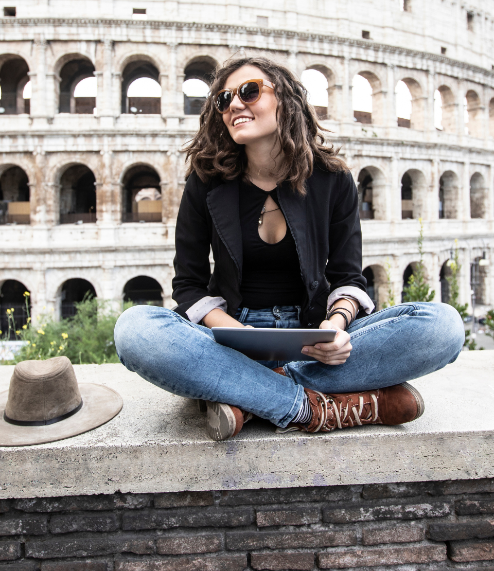 Young woman sitting with crossed legs and holding a tablet in front of the Colosseum