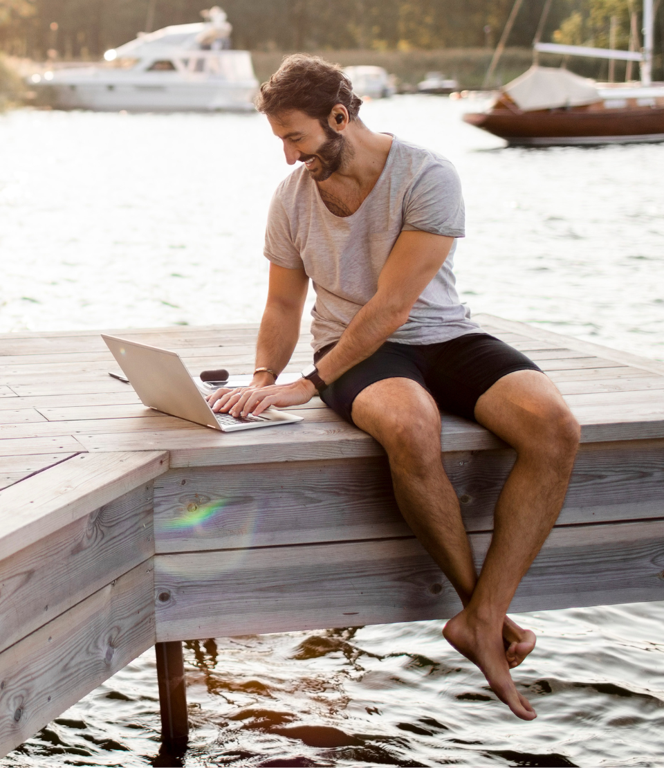 Young man sitting on a dock over water looking at his laptop and smiling