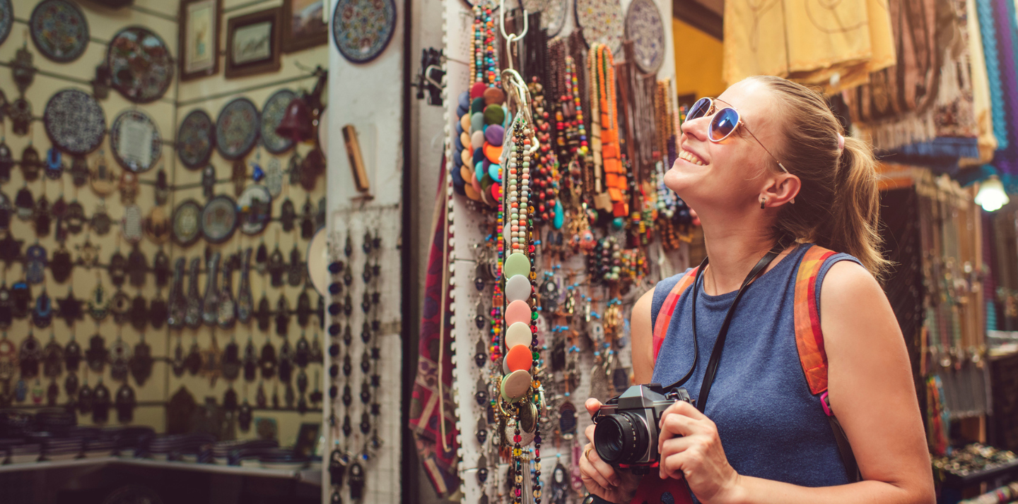 Young woman smiling looking at a store in a marketplace with her camera and solo travel insurance